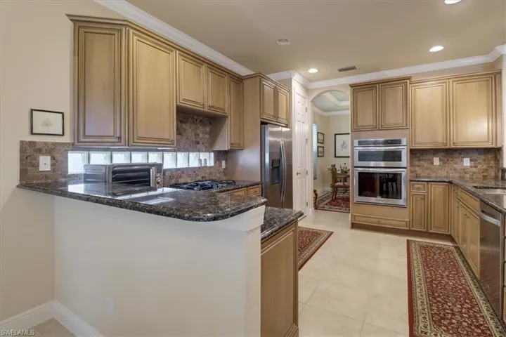 Kitchen featuring backsplash, appliances with stainless steel finishes, light tile patterned floors, dark stone countertops, and kitchen peninsula