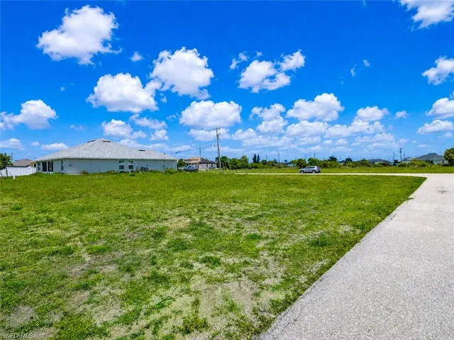 Expansive undeveloped lot featuring a concrete driveway, a flat terrain, and a distant single-story structure with multiple windows and a gray shingle roof