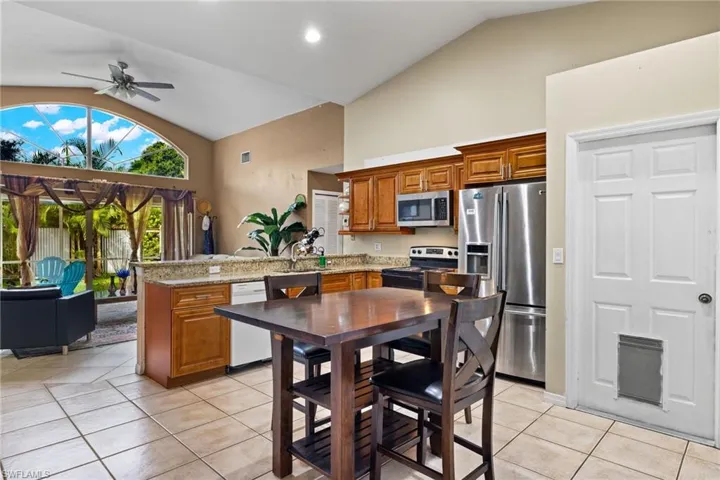 Kitchen with brown cabinetry, stainless steel appliances, light tile patterned flooring, light stone countertops, and high vaulted ceiling