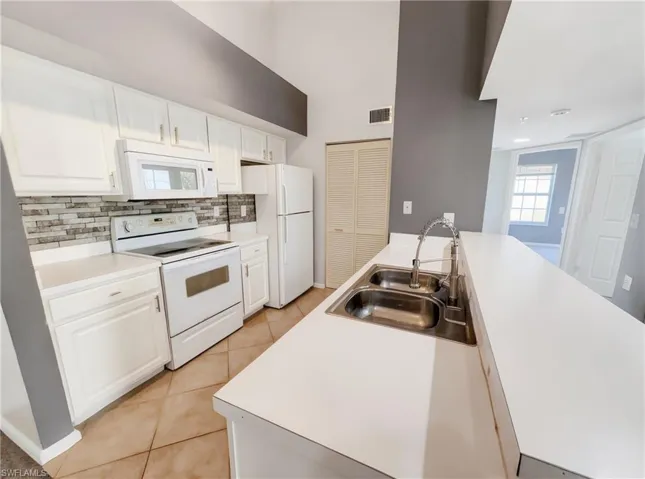 Kitchen featuring white cabinetry, white appliances, a stone-look tile backsplash, a double basin stainless steel sink, and tile flooring