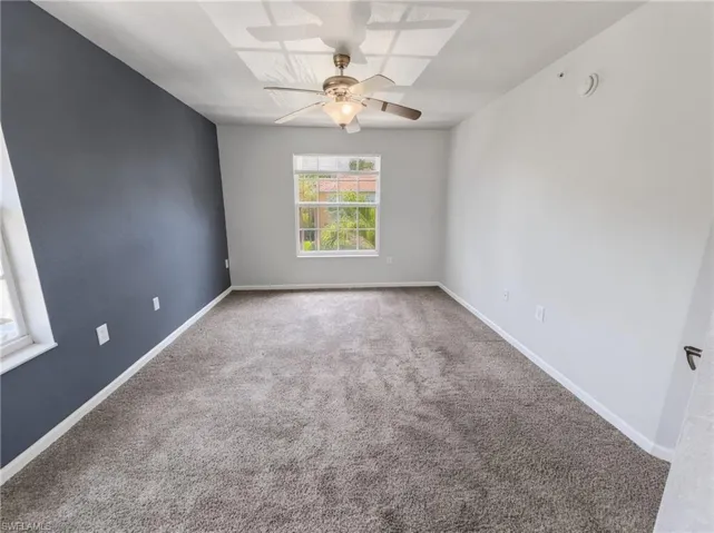 Spacious room featuring plush gray carpeting, a window offering natural light, a ceiling fan with integrated lighting, and a distinct accent wall