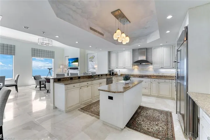 Kitchen with wall chimney range hood, a chandelier, a raised ceiling, light stone counters, and a center island
