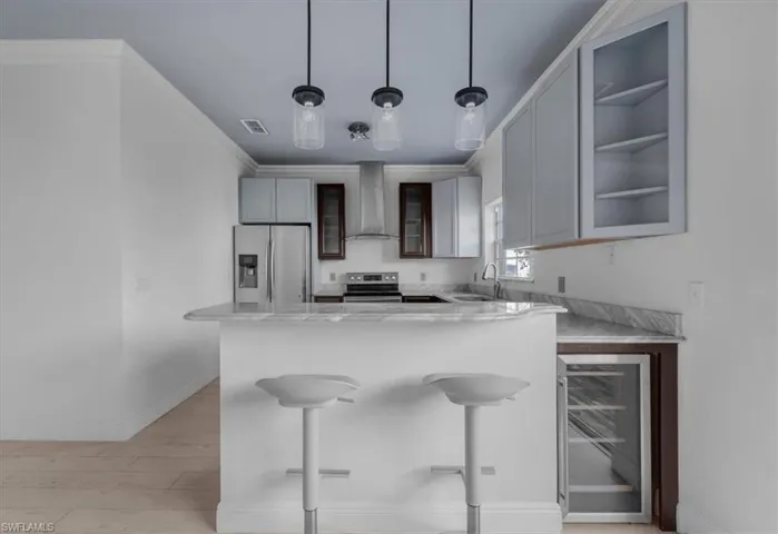 Kitchen featuring appliances with stainless steel finishes, beverage cooler, wall chimney range hood, decorative light fixtures, and light hardwood / wood-style floors