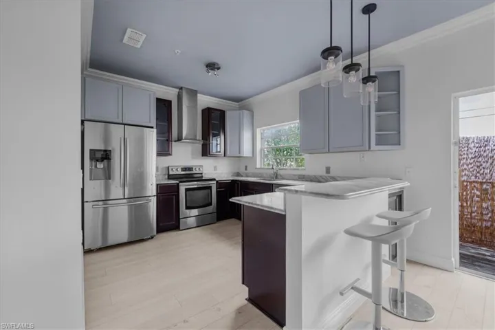 Kitchen with ornamental molding, a breakfast bar, stainless steel appliances, wall chimney range hood, and pendant lighting