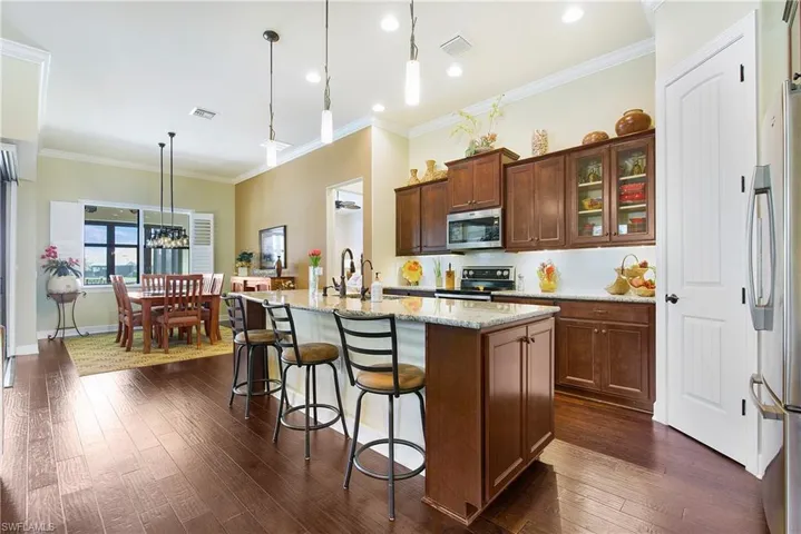 Kitchen with stainless steel appliances, dark wood-style flooring, light stone counters, a kitchen island with sink, and ornamental molding