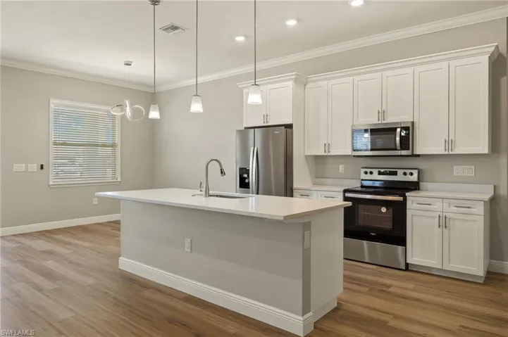 Kitchen featuring stainless steel appliances, white cabinets, an island with sink, hanging light fixtures, and crown molding