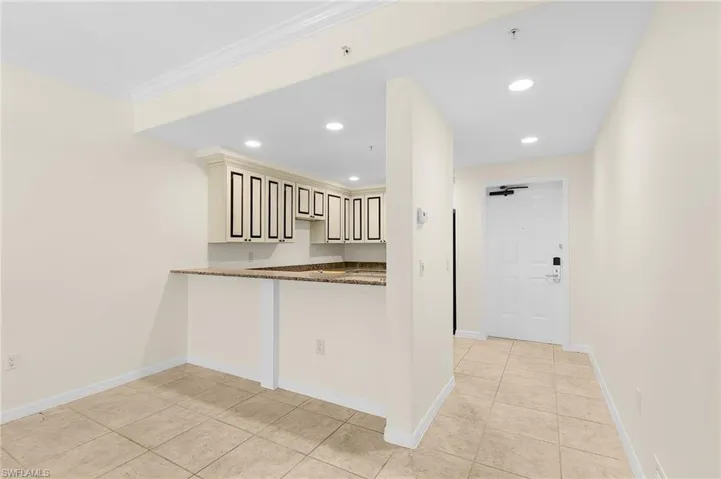 Kitchen and entry area featuring light-toned tile flooring, recessed lighting, crown molding, and a kitchen bar with a stone-finish countertop