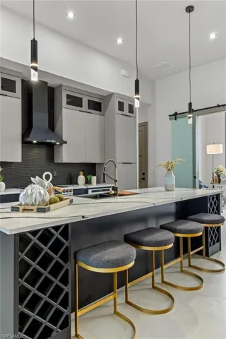 Kitchen with gray cabinets, decorative backsplash, hanging light fixtures, wall chimney exhaust hood, and a breakfast bar