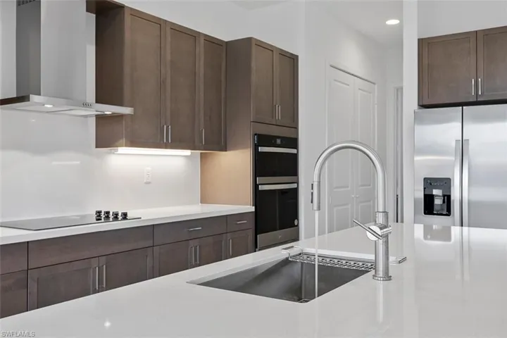 Kitchen featuring double oven, stainless steel refrigerator with ice dispenser, light countertops, wall chimney exhaust hood, and black electric cooktop