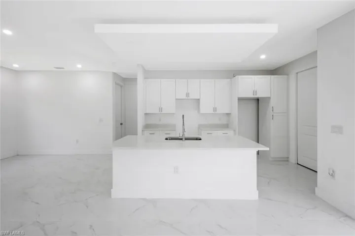 Kitchen with a kitchen island with sink, recessed lighting, white cabinetry, light marble finish floors, and light stone counters
