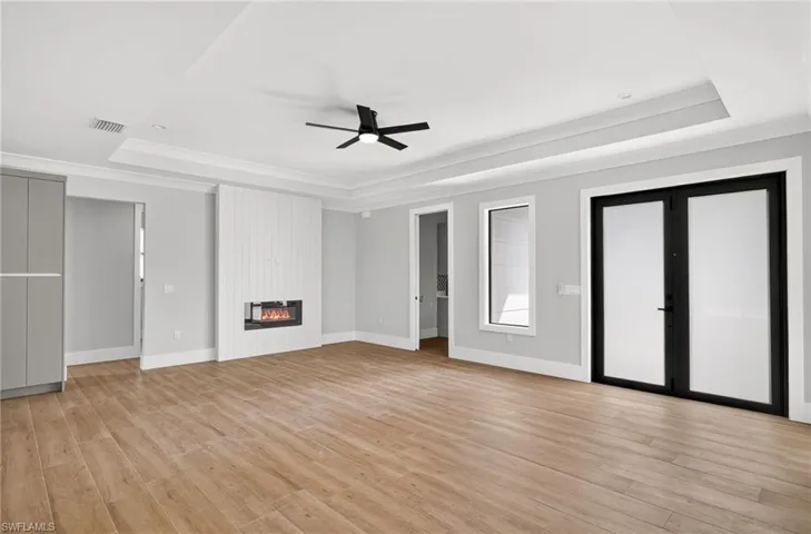 Tray ceiling featuring a central matte black ceiling fan, wood-finish flooring, white vertical plank fireplace surround, and black-framed French doors