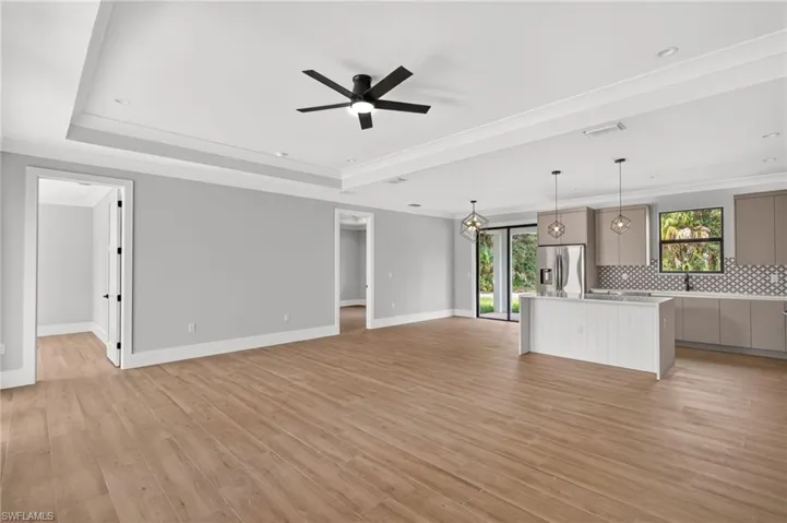 Open-concept interior featuring wood-finish flooring, a tray ceiling with a multi-blade ceiling fan, and a kitchen equipped with a central island, geometric pendant lighting, and a patterned tile backsplash