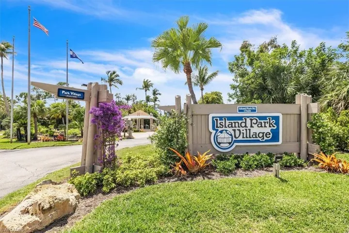 Island Park Village community entrance featuring a wooden welcome sign, lush tropical landscaping, and mature palm trees