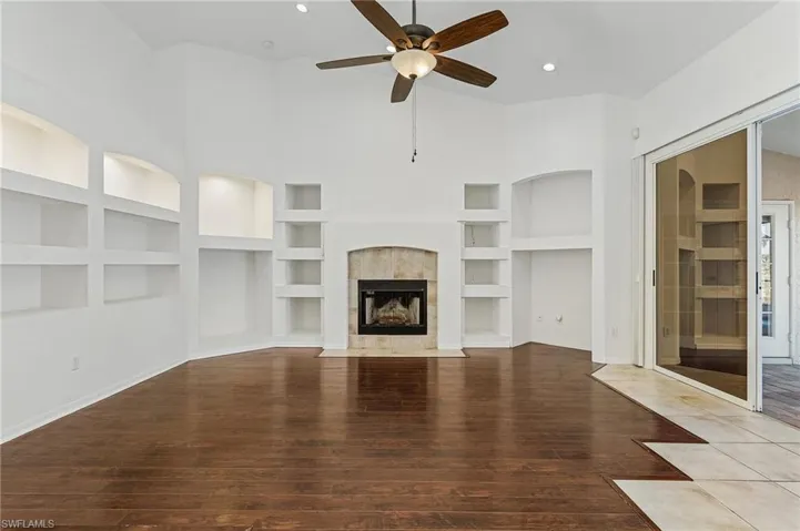 Unfurnished living room featuring a tiled fireplace, built in shelves, ceiling fan, wood-type flooring, and a towering ceiling