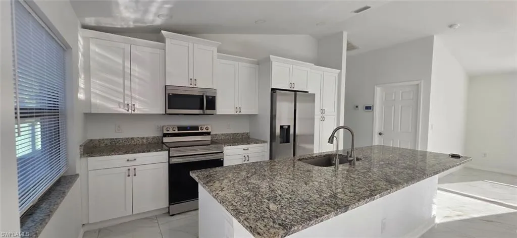 Kitchen with stainless steel appliances, white cabinets, dark stone countertops, vaulted ceiling, and a center island with sink