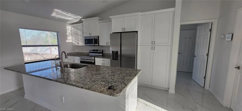 Kitchen with dark stone countertops, white cabinets, stainless steel appliances, light marble finish flooring, and vaulted ceiling