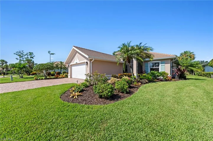 Charming single-story residence featuring a pristine manicured lawn, mature landscaping with tropical palms and flowering shrubs, brick paver driveway, and a two-car garage with white panel doors