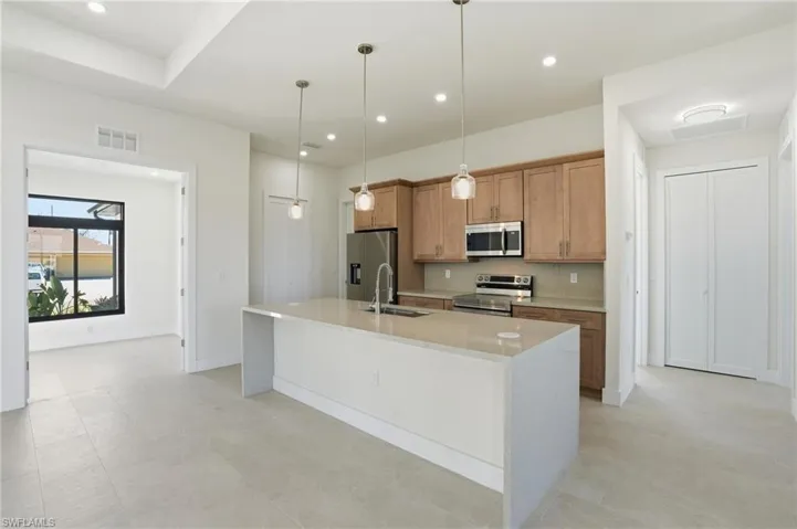 Kitchen with light stone countertops, pendant lighting, stainless steel appliances, a kitchen island with sink, and wood finish cabinets