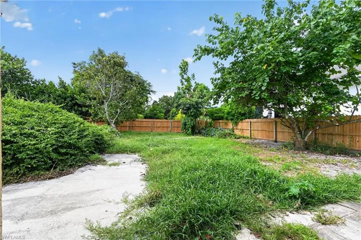 Spacious outdoor area featuring a wooden privacy fence, mature trees, dense shrubs, and a concrete pad