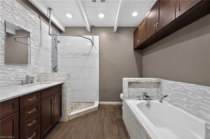 Bathroom featuring a walk-in shower with pebble tile floor, a soaking tub with tiled surround, wood-finish flooring, a vanity with a white countertop and wood cabinetry, and overhead storage cabinets