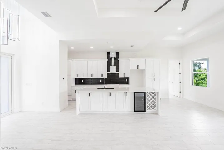 Kitchen featuring white cabinetry, wine cooler, wall chimney range hood, backsplash, and an island with sink