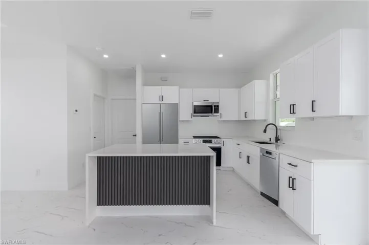 Contemporary kitchen featuring a center island with a dark fluted panel base, white flat-panel cabinetry with black hardware, stainless steel appliances, a black gooseneck faucet, and light-colored flooring