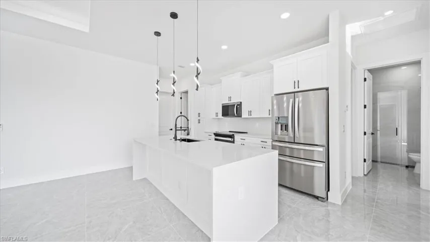 Kitchen with stainless steel appliances, hanging light fixtures, light stone counters, white cabinetry, and an island with sink