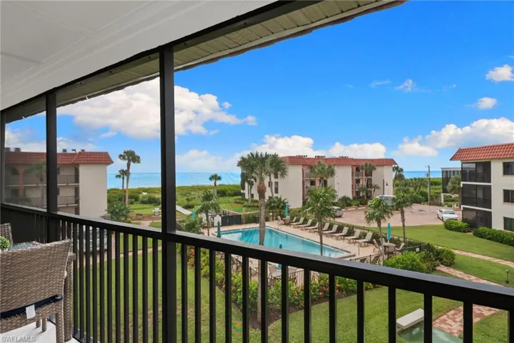 Screened balcony with black metal railings, overlooking a swimming pool, landscaped grounds, and beachfront