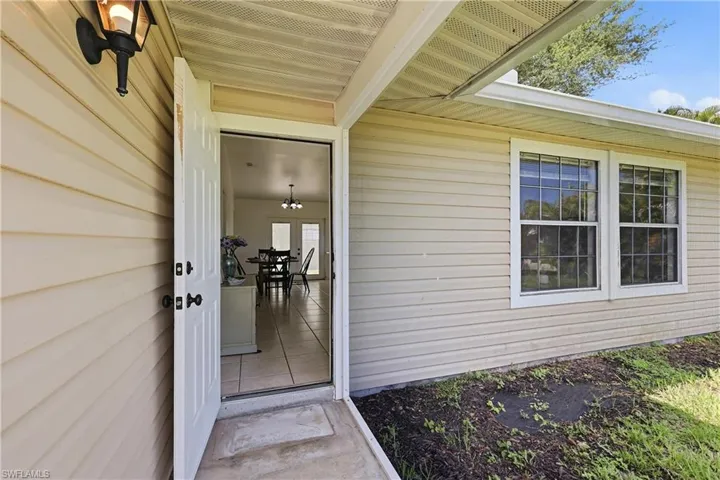 Cream-colored siding exterior featuring a white paneled front door, an exterior wall lantern, two double-hung windows with grilles, and a covered entryway