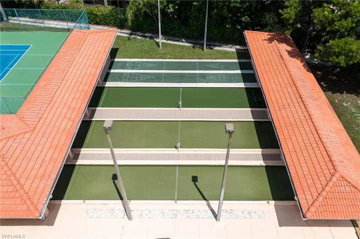 Multiple shuffleboard courts with green surfacing and white markings, bordered by two structures featuring terra cotta tile roofs