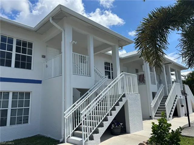 White stucco exterior with a blue accent band, featuring multiple entryways with white railings, a metal roof, and visible palm fronds