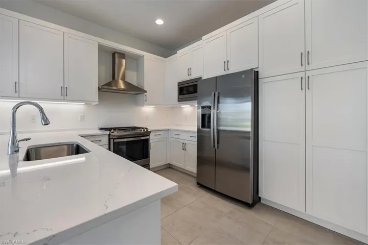 Kitchen with light stone countertops, stainless steel appliances, sink, wall chimney range hood, and white cabinets