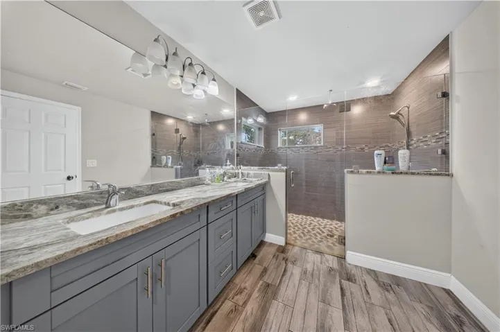 Double vanity with stone-finish countertop, walk-in shower featuring glass panel enclosure, horizontal wall tile, and wood-finish flooring
