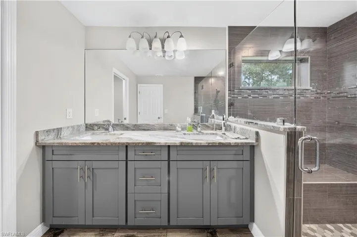 Double-sink vanity with stone countertop, gray shaker-style cabinetry, full-width mirror, and glass-enclosed shower with horizontal wall tile