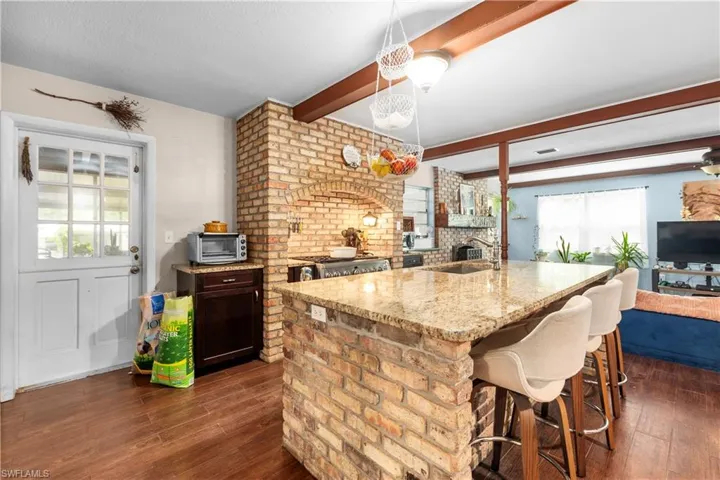 Kitchen with hanging light fixtures, a kitchen island with sink, light stone countertops, beam ceiling, and dark wood-type flooring