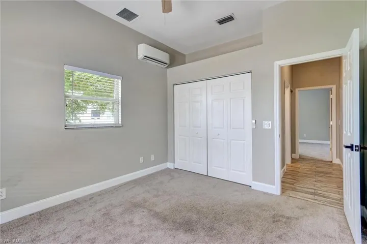 Carpeted interior space featuring a window with blinds, bi-fold closet doors, a wall-mounted air conditioning unit, and a ceiling fan
