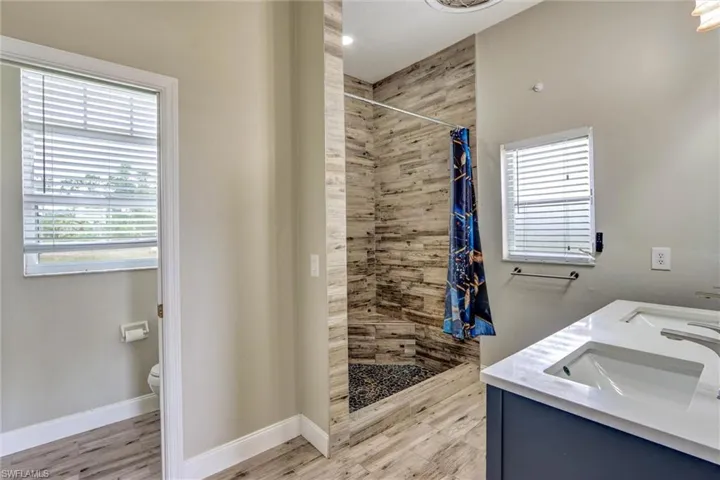 Bathroom featuring a walk-in shower with wood-finish tile surround and pebble floor, dual vanity with white countertops, and wood-finish flooring