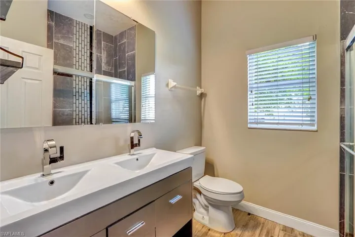 Bathroom featuring a dual-sink vanity with modern fixtures, large wall-mounted mirror, wood-finish flooring, and a tiled shower enclosure