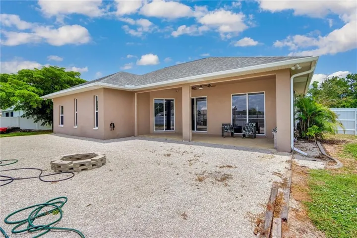 Rear exterior featuring a covered patio with recessed lighting and a ceiling fan, multiple sliding glass doors, a gravel-covered yard with a built-in fire pit, and a white privacy fence