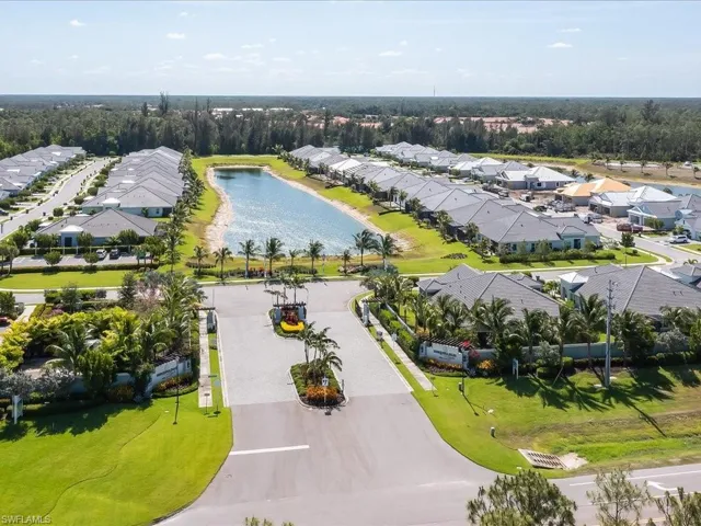 Community entrance with landscaped grounds, a paved road, and a central water feature bordered by housing