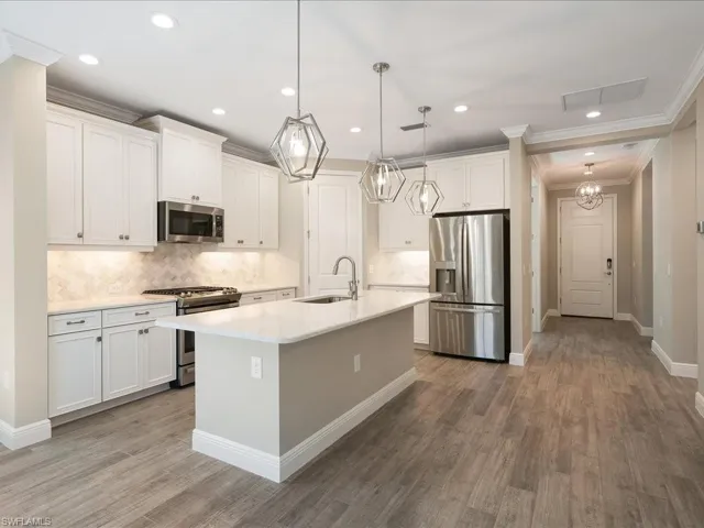 White shaker cabinetry, stainless steel appliances, stone tile backsplash, wood-finish flooring, and a central island with a sink