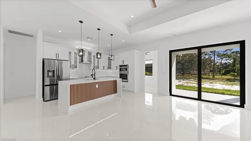 Kitchen with glass insert cabinets, stainless steel refrigerator with ice dispenser, an island with sink, white cabinets, and hanging light fixtures