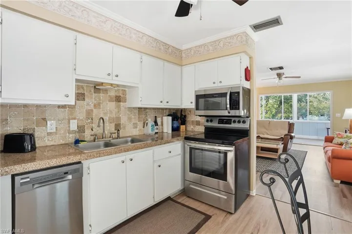 Kitchen featuring stainless steel appliances, tile countertops, backsplash, white cabinets, and crown molding - Virtually Edited Image