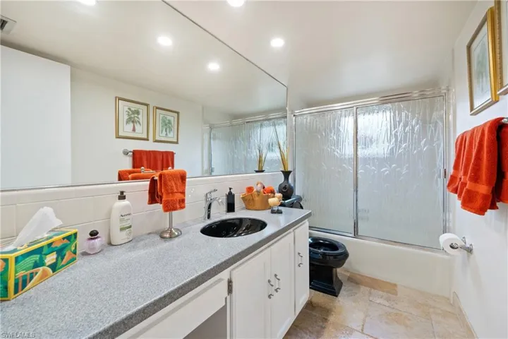 Full renovated bath with double vanity, combined bath/shower with glass door, light stone finish floors, one piece Silestone countertop and recessed lighting