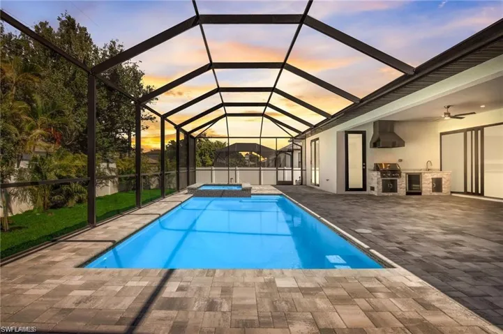 Pool at dusk with glass enclosure, grilling area, an outdoor kitchen, an in ground hot tub, and a patio