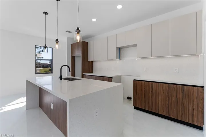 Kitchen with sink, backsplash, light stone counters, an island with sink, and decorative light fixtures