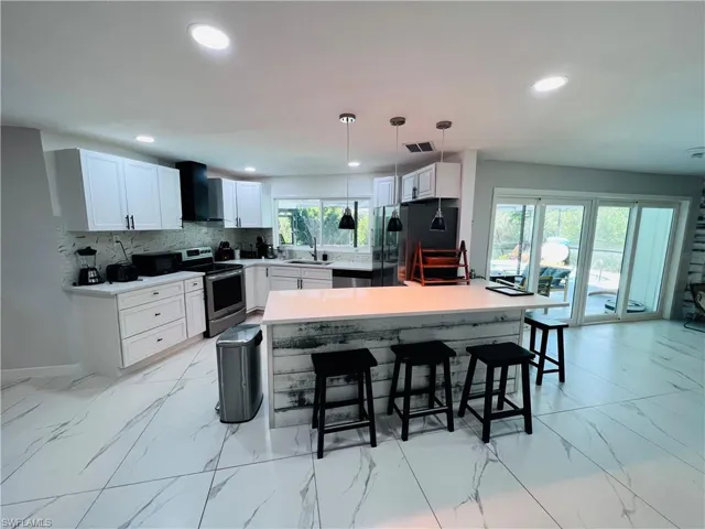 Modern kitchen with white cabinetry and stainless steel appliances, featuring a large center island with a light-toned countertop, white marbled-look tile flooring, and recessed ceiling lighting