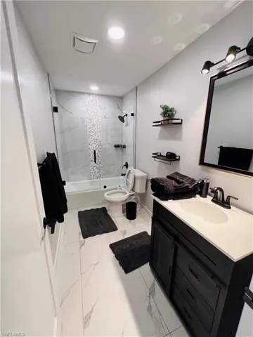 White tile flooring with subtle veining, dark wood-finish vanity with a white countertop, wall-mounted mirror, and a tub-shower combination with glass enclosure