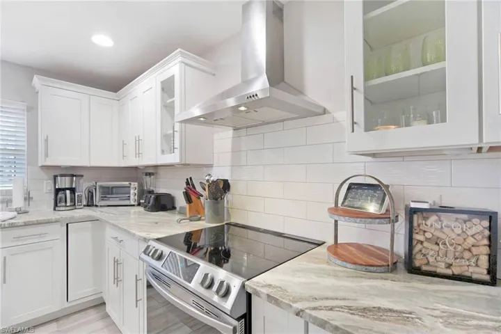 Kitchen featuring glass insert cabinets, stainless steel electric range oven, wall chimney range hood, light stone countertops, and white cabinetry
