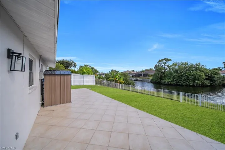 Rear exterior featuring a tiled patio, manicured lawn, perimeter fencing, and waterfront frontage with a metal storage unit